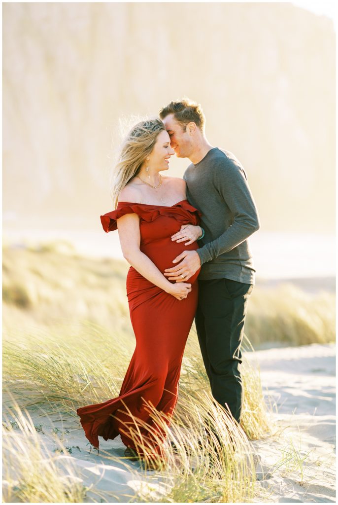 Couple laughing with woman in long red dress at beach maternity photo shoot
