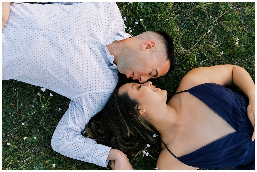 Couple laying on the grass head to head with eyes closed holding hands. 