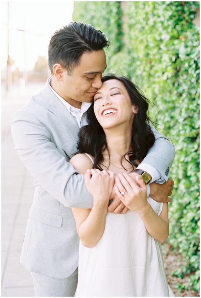 Man hugging fiance from behind around the shoulders while she smiles. 