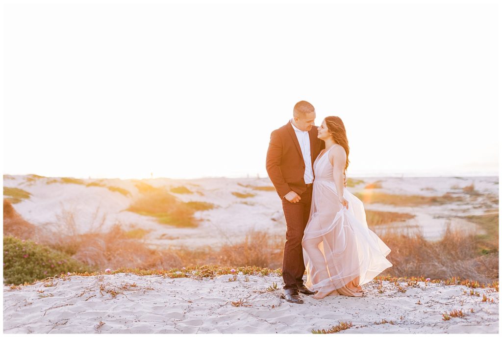 Woman in a sheer floor length dress with fiance in a suit at sunset in the sand. 
