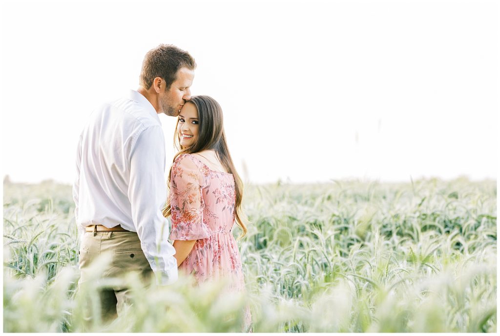 Engaged couple in wheat field in spring. Woman in pink floral dress while fiance kisses her forehead