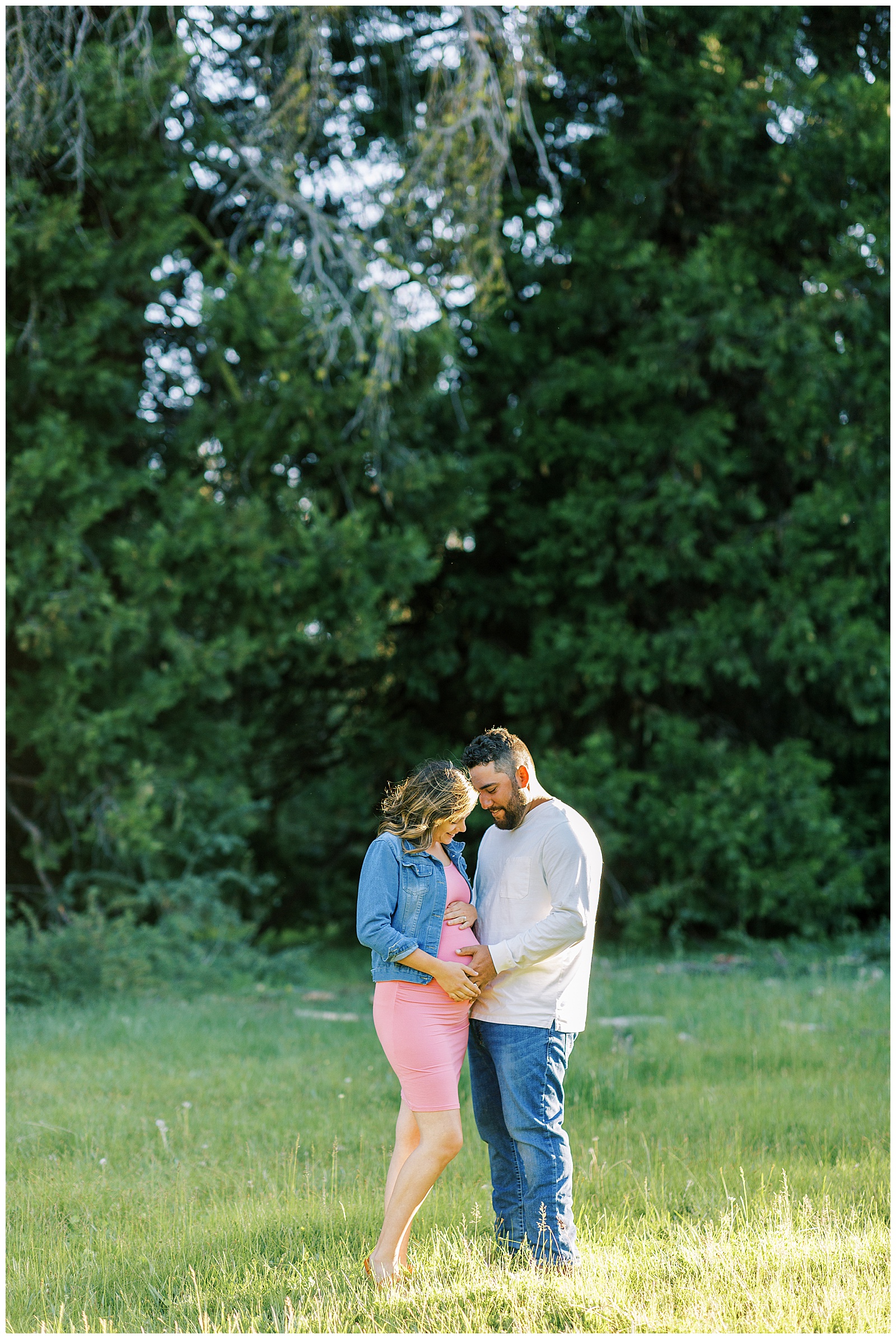 husband and wife looking at pregnant belly in pink dress standing in grassy meadow golden sunlight