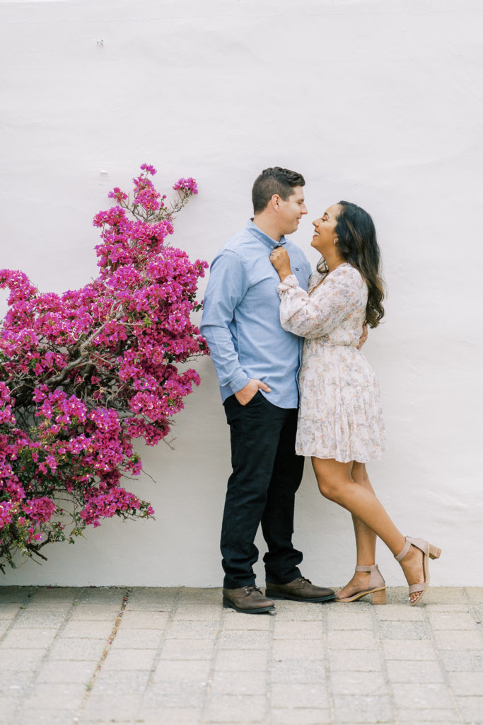 engaged couple standing in front of a white stone wall with a pink bougainvillea for engagement photos in monterey