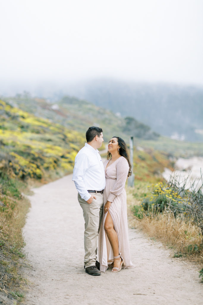 couple standing on a dirt path along carmel beaches while she wear a maxi dress from baltic born clothing and he wears khakis and a white button down shirt 