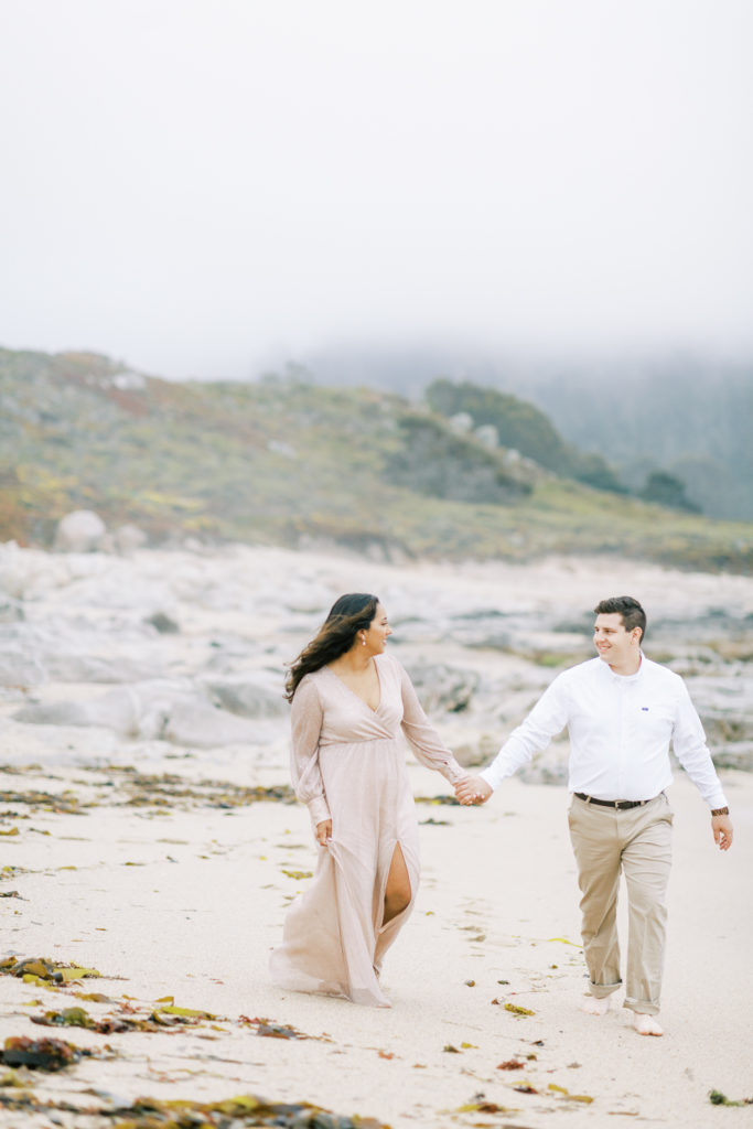couple holding hands and walking along a foggy beach in carmel for engagement photos