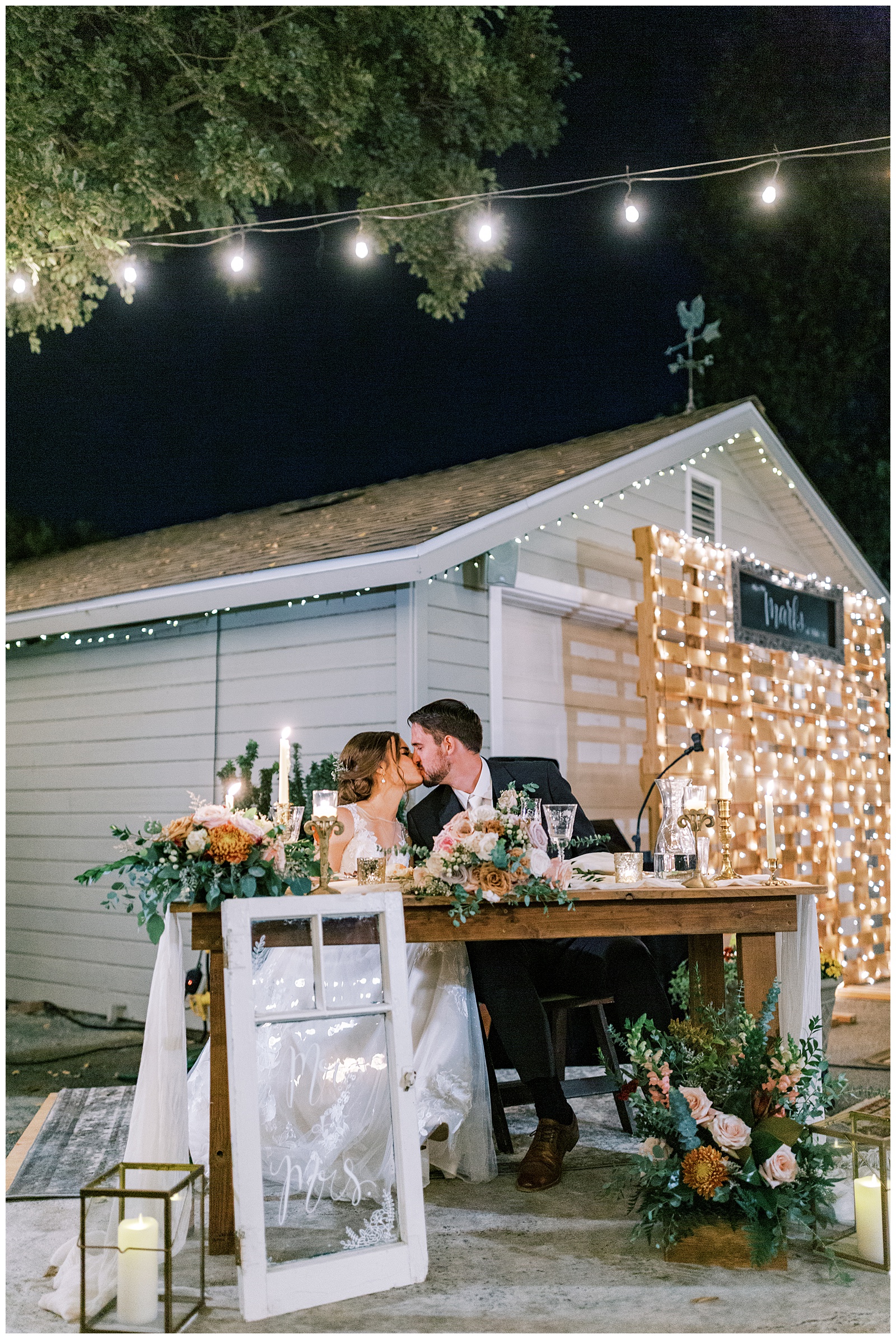bride and groom kissing at sweetheart table decorated with flowers and candles with white christmas lights in background