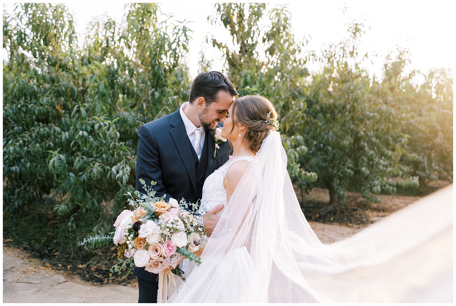 bride and groom embracing foreheads touching veil sweeping out golden sunlight shining in corner blush bouquet
