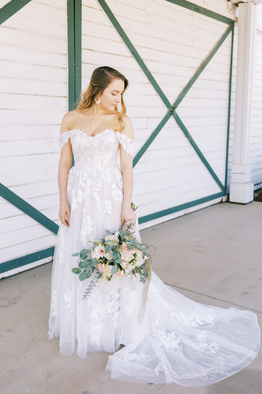 bride in front of white barn doors holding pink, white and eucalyptus wedding bouquet off the shoulder floral lace wedding dress at springville ranch