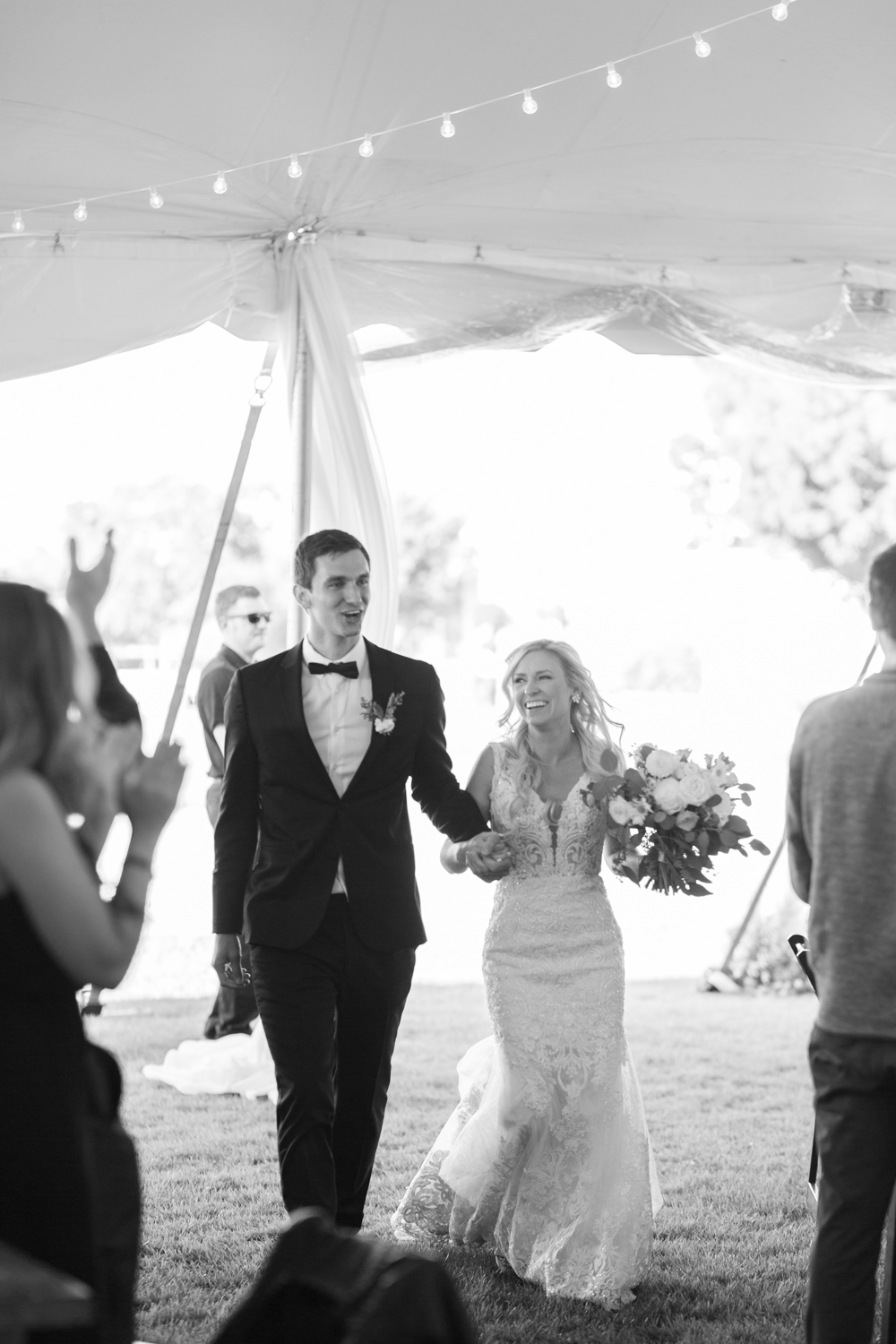 black and white photo bride and groom holding hands walking into wedding reception tent smiling guests clapping in background