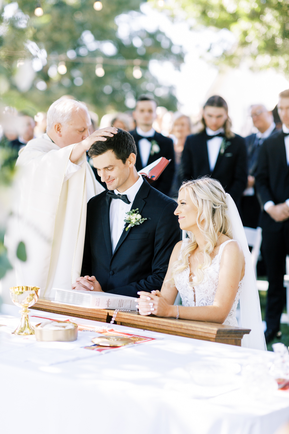 catholic priest blessing bride and groom during backyard wedding ceremony outdoors