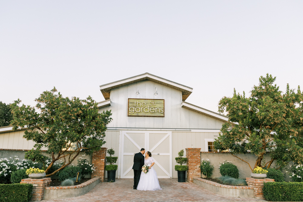 bride and groom standing in front of metal barn wedding venue the gardens tulare