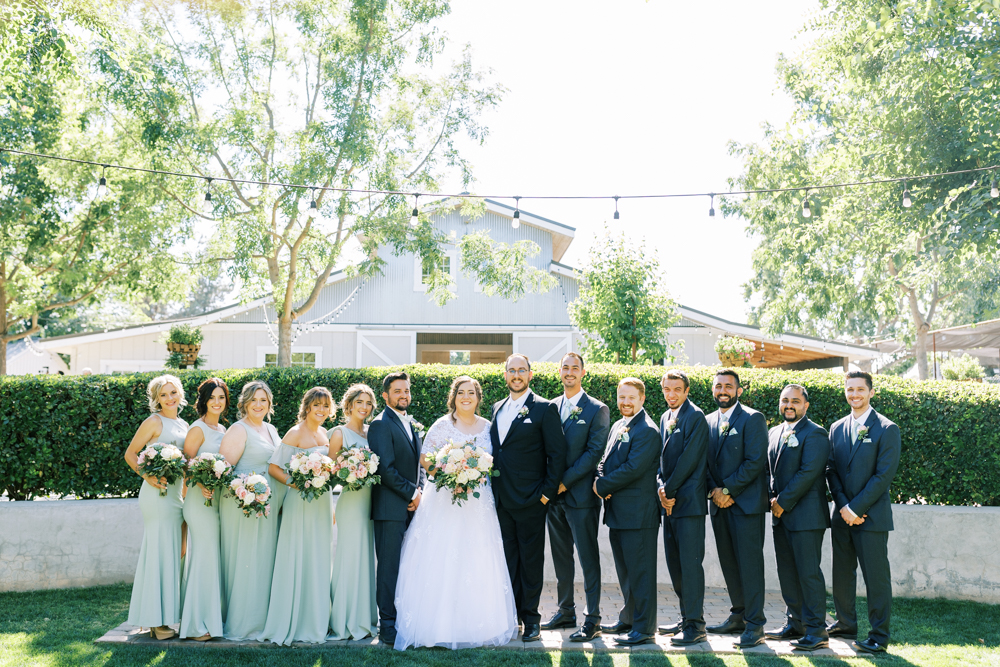 wedding party in sage green dresses and dark grey suits at barn wedding venue the gardens venue tulare california