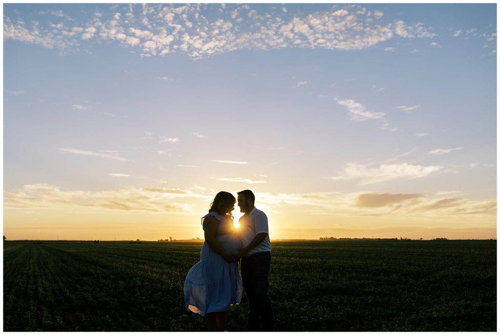 silhouette of pregnant couple in open field at sunset with sunlight glowing behind them 