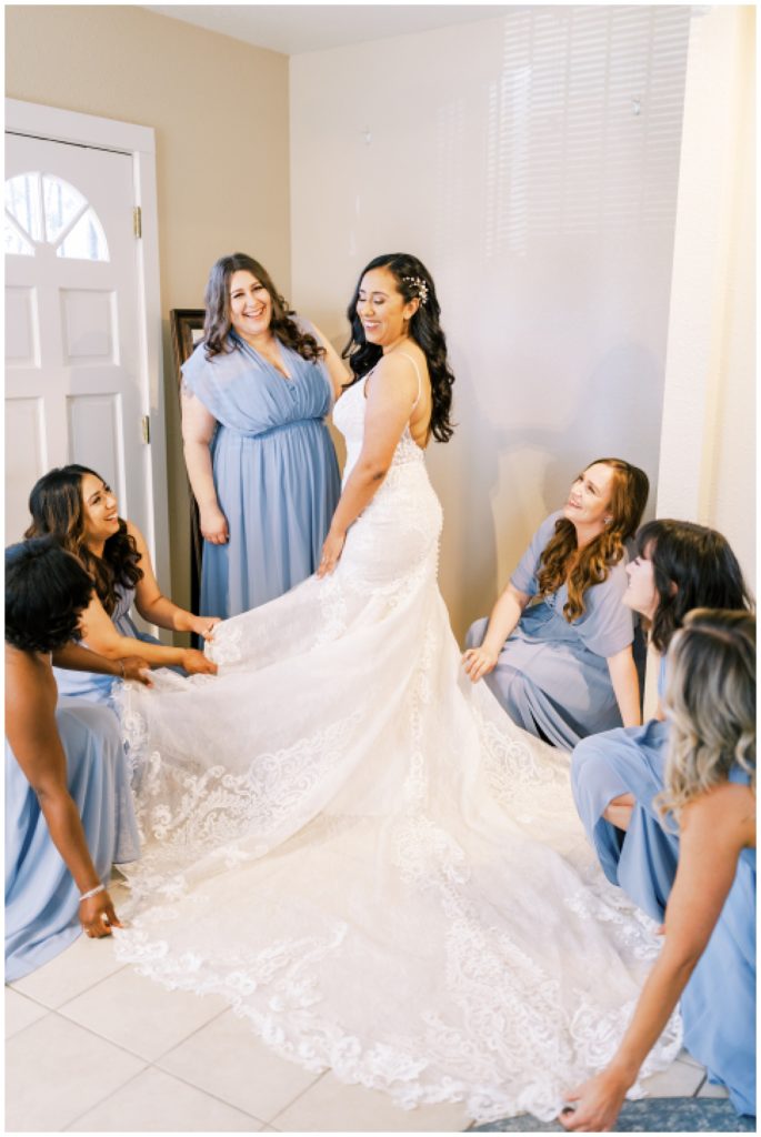 smiling bride putting on wedding dress with bridesmaids fanning out lace train