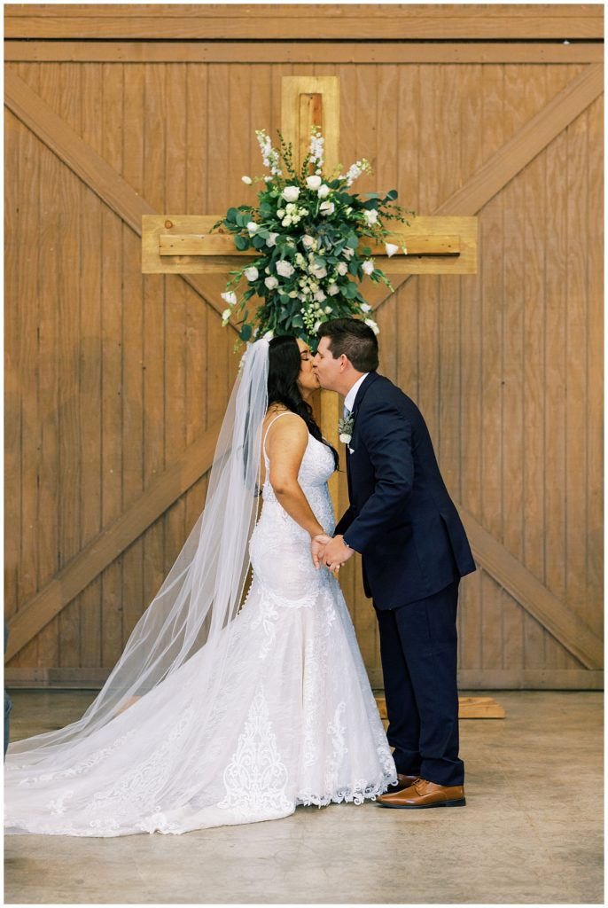 bride and groom first married kiss in front of wooden cross indoor wedding ceremony