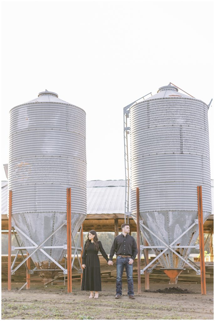 couple wearing black holding hands standing in front of metal silos