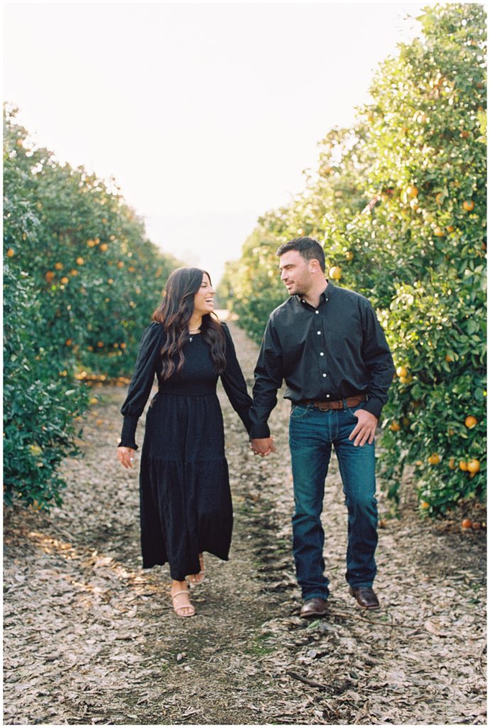 man and woman holding hands walking through orange grove smiling and laughing