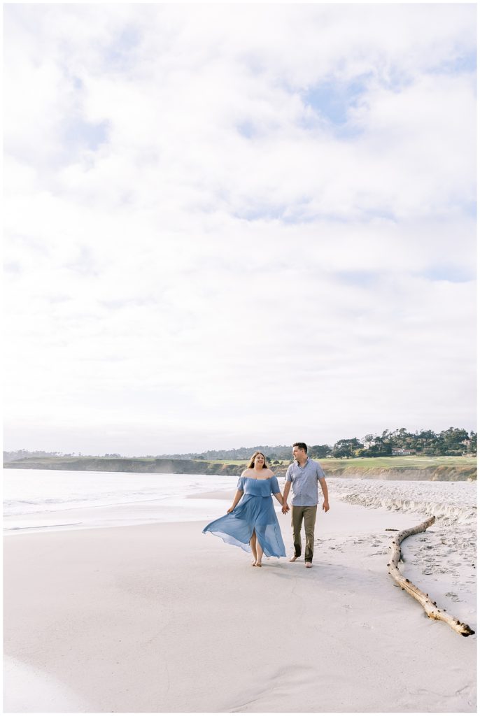 woman in blue chiffon dress holding hands with fiance walking on carmel beach engagement photos